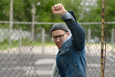 A man stands in front of a chain-link fence, raising his fist in a gesture of solidarity. He wears a...