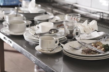 Dirty cups and plates with leftovers and silverware placed on metal table ready for dish washing in...