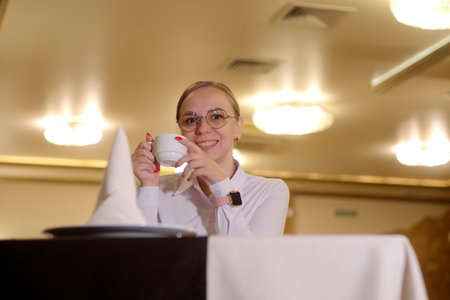 Woman with cup of coffee. Young female in casual clothes and eyeglasses sitting at table with cup of...