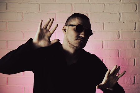 Stylish man poses confidently against a pink-lit brick wall while wearing sunglasses and a dark hoodie indoors at night.の写真素材