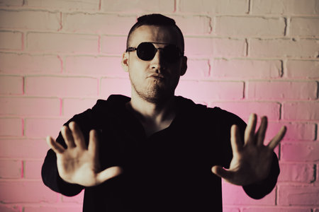 Stylish man poses confidently against a pink-lit brick wall while wearing sunglasses and a dark hoodie indoors at night.の写真素材