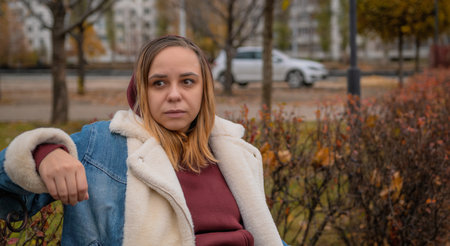 A woman sitting thoughtfully on a park bench surrounded by autumn foliage in an urban setting during late afternoon.の写真素材