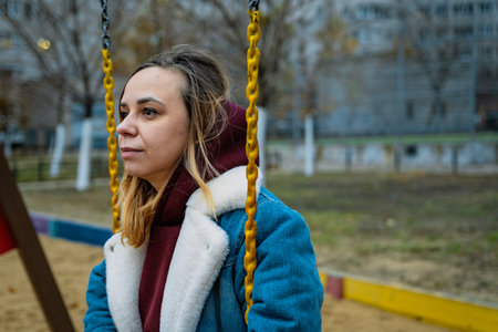 A woman enjoys swinging at a playground surrounded by trees and apartment buildings on a cool autumn day.の写真素材