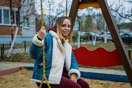A woman enjoys swinging at a playground surrounded by trees and apartment buildings on a cool autumn...