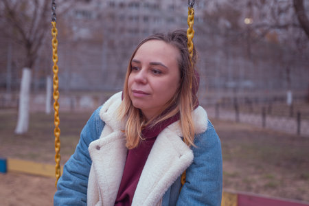 A woman enjoys swinging at a playground surrounded by trees and apartment buildings on a cool autumn...