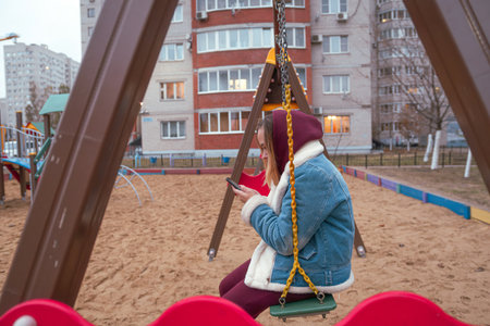 Young woman in a hooded jacket uses her smartphone while sitting on a swing at a playground during...