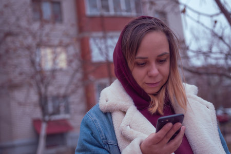 A young woman checks her smartphone while standing outside a residential building in early evening...