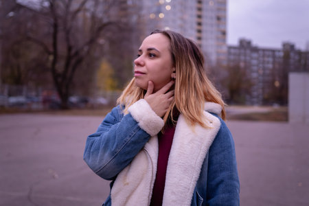 Young woman in casual attire stands in an urban park during early evening, surrounded by tall buildings and autumn trees.の写真素材