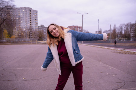 Young woman in casual attire stands in an urban park during early evening, surrounded by tall...