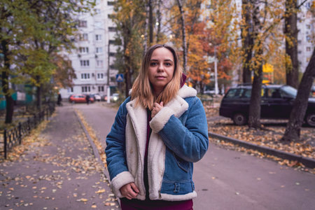 A woman stands thoughtfully in an urban setting during autumn, surrounded by trees and parked cars...
