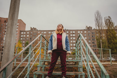 A confident woman stands on rusted stairs in a dilapidated urban area during a cloudy autumn day.