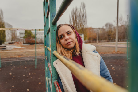 Young woman in a hoodie and coat poses at a playground during autumn in a city park, surrounded by colorful trees and apartment buildings.の写真素材