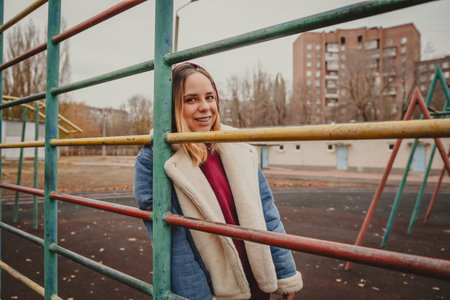 Young woman in a hoodie and coat poses at a playground during autumn in a city park, surrounded by...