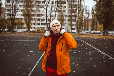 Man in orange jacket stands in urban park surrounded by bare trees and large apartment building...