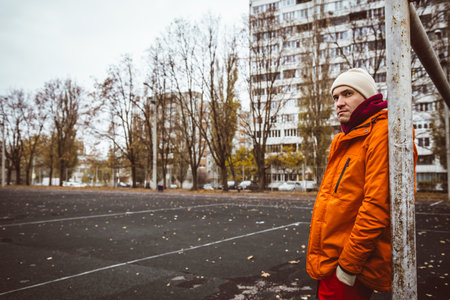 Young man in an orange jacket stands pensively by a post on an empty sports field during a chilly...