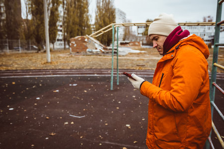 Man in orange jacket checks phone in empty playground during autumn afternoon