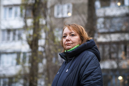 Woman in winter coat stands outside apartment building in urban setting during early evening hours.