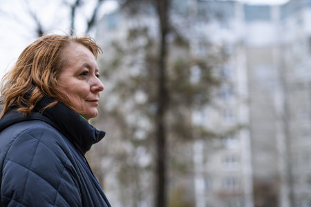 Woman in winter coat stands outside apartment building in urban setting during early evening hours.