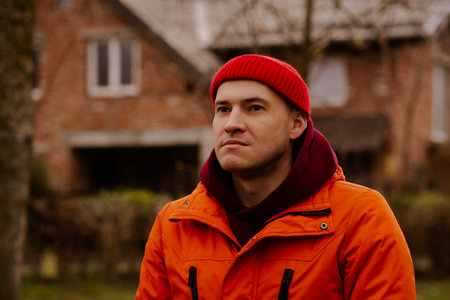 Young Man in Bright Orange Jacket and Red Beanie Standing Outdoors in a City During Winter Season.