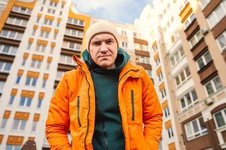 Man Wearing an Orange Jacket Standing in Front of Modern Apartment Buildings on a Sunny Day.の写真素材