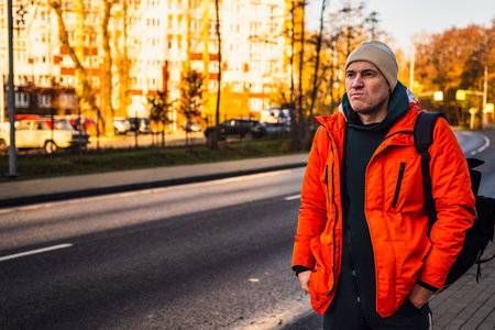 Man Wearing an Orange Jacket Stands by the Roadside During Sunset in an Urban Area.の写真素材