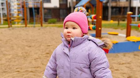 Cute Toddler in Purple Coat and Pink Hat Enjoys Playground on a Chilly Day at Outdoor Parkの写真素材