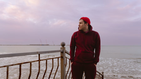 Young Man Stands by the Seaside in a Red Hoodie While Gazing at the Horizon During Sunsetの写真素材