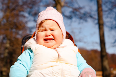 A young child sits in a stroller outdoors making an upset face and expressing sadness The setting...