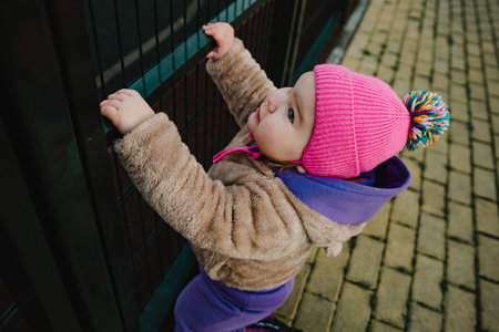 A young child with a serious expression reaches out to a metal fence wearing a cozy beige jacket and...