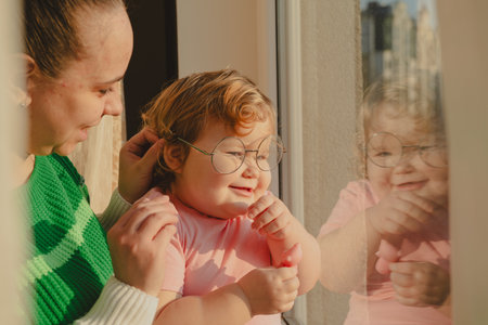 A young child smiles joyfully as she adjusts her glasses while sitting by a window She is held by a...