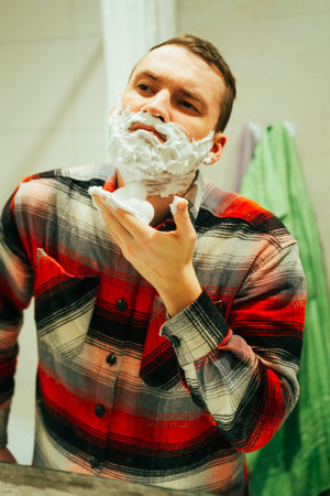 In a well-lit bathroom a man is focused on shaving as he applies shaving cream to his face He looks into the mirror while ensuring a clean shave.のeditorial素材