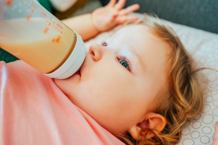 A young child with curly hair is calmly drinking from a bottle while lying on a cozy blanket...