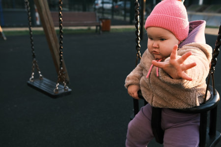 Child is having fun swinging in a park while wearing a pink hat and cozy outfit The bright morning light adds a cheerful atmosphere to the playful moment.のeditorial素材