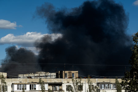 Thick black smoke rises from damaged residential buildings in an urban setting. The sky is clear,...