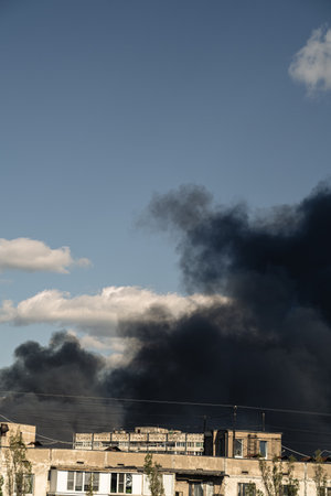 Thick black smoke rises from damaged residential buildings in an urban setting. The sky is clear,...
