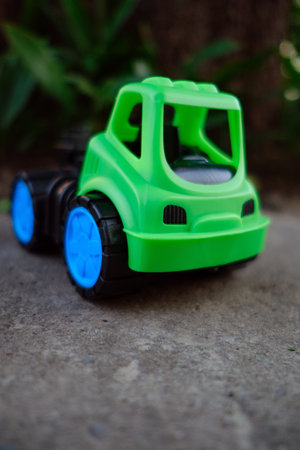 A vibrant green toy truck with blue wheels rests on a gravel surface. Sunlight shines gently,...