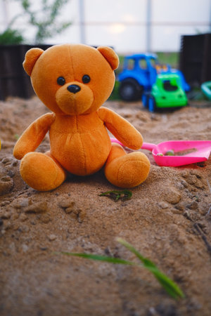 A soft orange teddy bear is placed in a sandbox with vibrant plastic toys scattered nearby The sand glistens under bright sunlight creating a joyful atmosphere for play.の写真素材