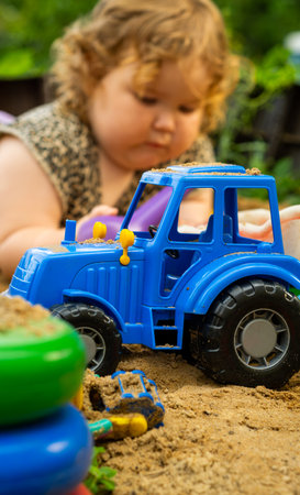 Blue toy tractor moving through sand with children playing nearby at a sunny outdoor location.の写真素材