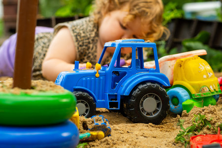 Blue toy tractor moving through sand with children playing nearby at a sunny outdoor location.の写真素材