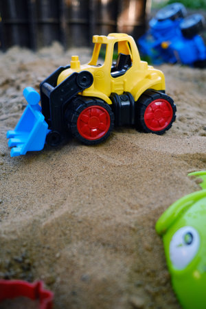 A bright yellow bulldozer toy digs in the sand at a playground filled with colorful plastic toys The scene captures fun outdoor playtime under clear blue skies and sunlight.の写真素材