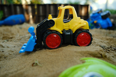 A bright yellow bulldozer toy digs in the sand at a playground filled with colorful plastic toys The scene captures fun outdoor playtime under clear blue skies and sunlight.の写真素材