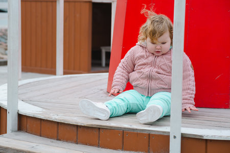 A young child dressed in a cozy sweater and light pants sits on a wooden structure at an outdoor playground The wind gently tousles her hair as she explores her surroundings.の写真素材