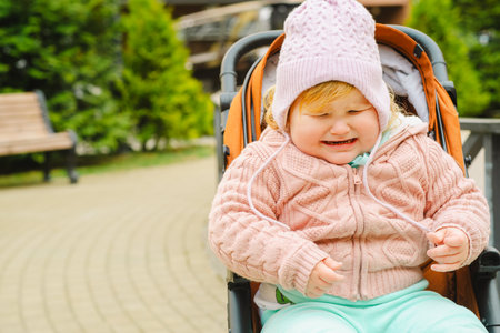 A young child in a cozy sweater and hat sits in a stroller while crying in a vibrant park Lush trees and pathways surround the area providing a lively atmosphere for outdoor activitiesの写真素材