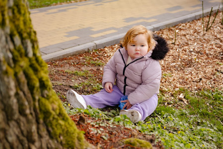 A young child sits on the ground in a park exploring the grass and leaves while wearing a warm coat The tree nearby adds a natural touch to the setting.の写真素材