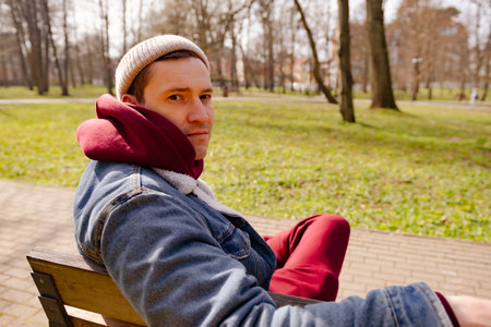 Man wearing a beanie and hoodie relaxes on a bench in a park on a sunny spring day The vibrant green grass and trees create a tranquil atmosphere for reflection.の写真素材