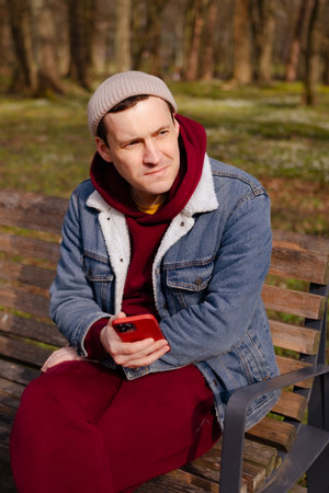 A young man dressed in a cozy red hoodie and denim jacket sits on a wooden bench in a park absorbed in his smartphone surrounded by trees on a bright day.の写真素材
