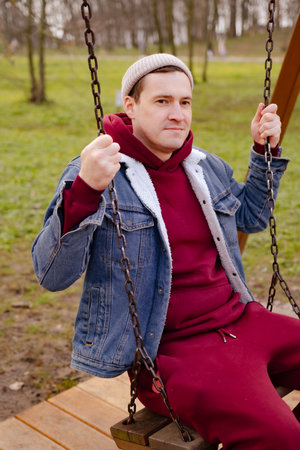 A young man sits calmly on a swing in a park surrounded by trees as autumn leaves cover the ground. He wears a denim jacket over a red hoodie, creating a relaxed vibe.の写真素材