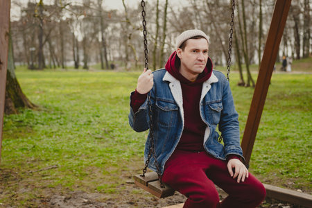 A young man sits calmly on a swing in a park surrounded by trees as autumn leaves cover the ground He wears a denim jacket over a red hoodie creating a relaxed vibe.の写真素材