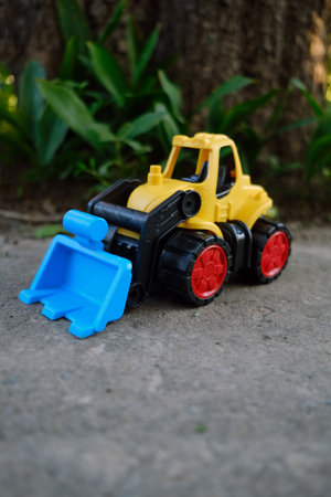 Bright Yellow Toy Construction Vehicle With Blue Scoop Resting on Dirt Near Greenery in Sunny...