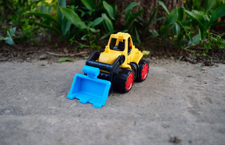 Bright Yellow Toy Construction Vehicle With Blue Scoop Resting on Dirt Near Greenery in Sunny...